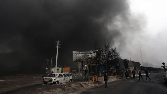 A smoky charred street with cars parked and people standing looking on