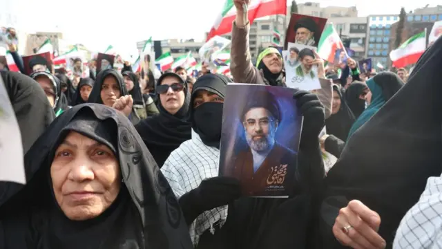 A woman holds a small poster with an image of Mojtaba Khamenei in a public gathering celebrating his appointment as Supreme Leader.