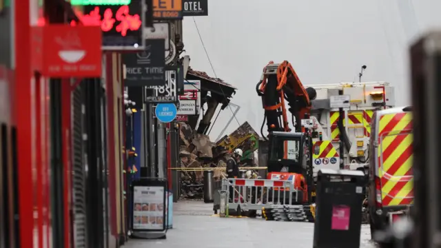 Close-up of rubble that has spilled onto the pavement. There are also shop fronts and emergency vehicles on the image.