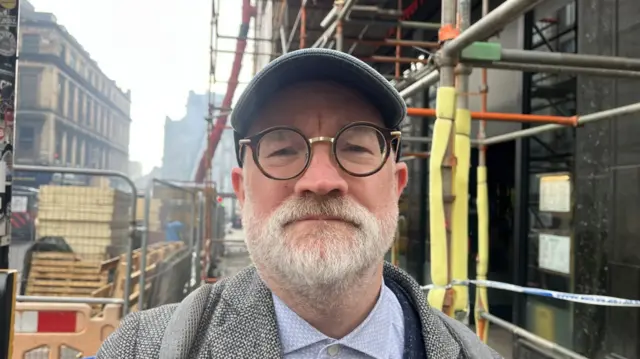 A man with a grey beard and round black glasses wearing a hat. he is standing under some scaffolding.