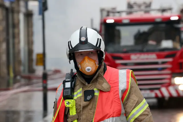 Firefighter wearing protective gear including an orange face mask and a high‑visibility vest standing on a wet street in Glasgow, with a fire engine and emergency scene in the background.