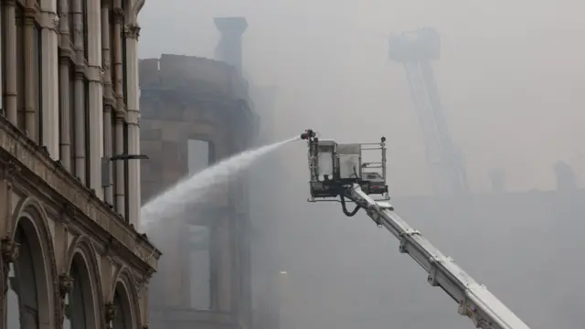 Aerial firefighting platform spraying water onto a smoke‑filled building in Glasgow, with heavy haze obscuring surrounding structures.