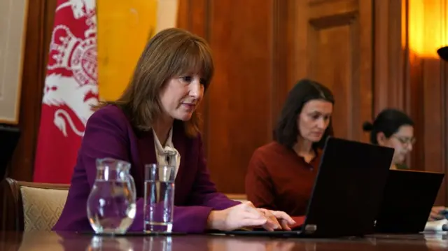 Reeves sitting at a table looking at a laptop while she types on it. Two women sit next to her doing the same. There's a jug and glass of water next to her.