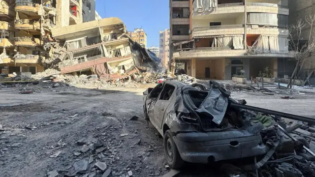 A car appears burnt and badly damaged, with a partially collapsed building behind