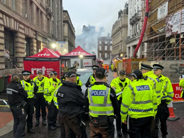 Police officers on the street with smoke surrounding the burnt-out building in the background