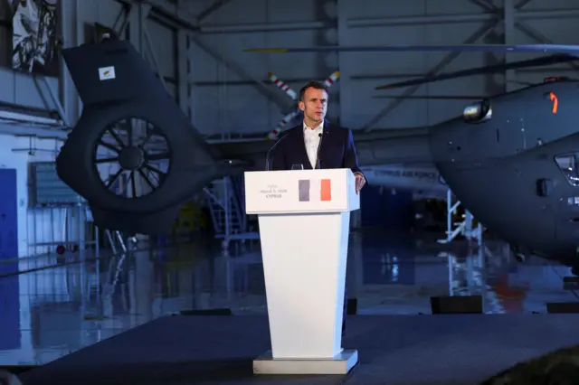 Emmanuel Macron standing at press conference podium taking place in a hangar. There's the tail end of what appears to be a military helicopter behind him.