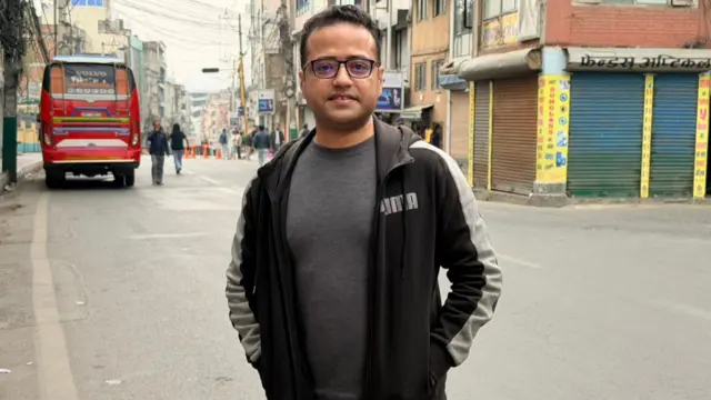 Raghunath Neupane smiles at the camera. He wears a pair of glasses and puts his hands in the pockets of the black sports jacket he is wearing