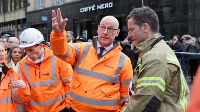 John Swinney, wearing a hi-vis ScotRail jacket, gestures upwards while talking to a firefighter.  There are also ScotRail officials in hi-vis jackets