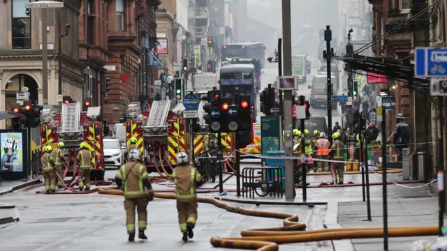 Fire engines and firefighters on a road with traffic ahead. There is a large hose across the road.