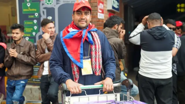 Raju Kumar Lamichhane smiles at the camera. He wears a red hat with a Nepali flag wrapped around his neck.
