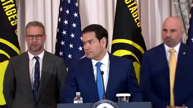 Rubio standing at a lectern looking to the side. Two men stand behind him, one on either side.