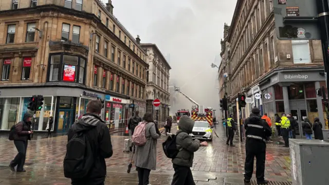 People on a street looking at firefighters on a high ladder surrounded by smoke