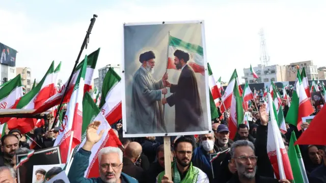 A man holds a placard appearing to show Ali Khamenei passing a flag onto this son Mojtaba in a public gathering celebrating the appointment of the new Iranian leader.