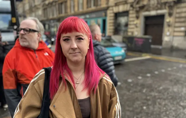 Courtney MacKinnon, who has bright pink hair shoulder-length hair with a fringe, is wearing a brown jacket. She is standing on a Glasgow street with pedestrians, vehicles and Victorian sandstone buildings in the background.