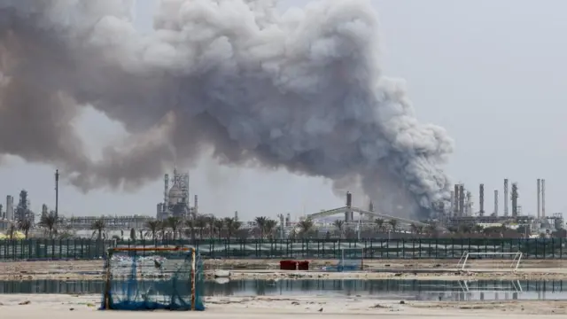 A large grey smoke plume rises over an oil refinery.