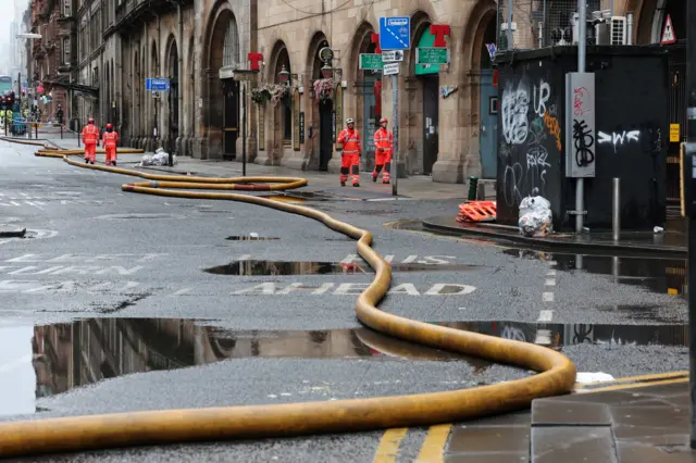 Fire hoses stretched across a wet roadway on Hope Street in Glasgow, with emergency workers in high‑visibility clothing standing near historic stone buildings and traffic signs.