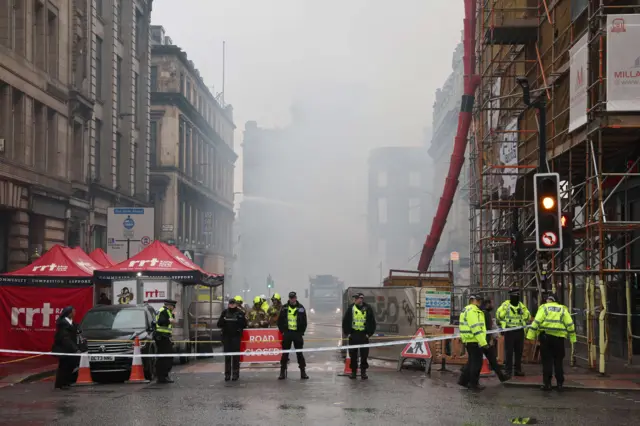 Police officers and emergency workers stand behind cordons on Renfield Street, Glasgow. The air is filled with heavy smoke, with surrounding buildings, scaffolding, tents, and traffic lights visible during an active incident response. Rapid Relief Team tents and a trailer are erected in front of the bus gate sign.