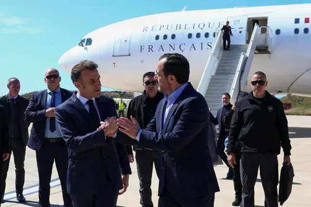 Macron shaking hands with Christodoulides on tarmac of runway. Behind them are men, who appear to be security officers, wearing dark clothing, and a plane with the word "Francais" on it and steps out descending to the tarmac.