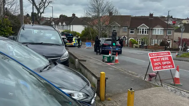 Armed police are standing next to two police cars by a temporary traffic light.