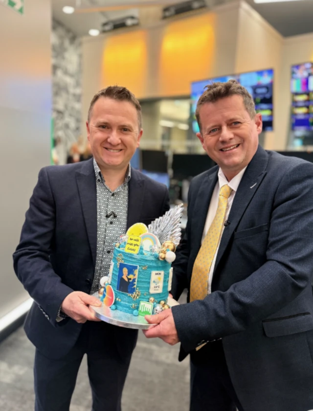 Simon King and Matt Taylor carrying a blue cake with weather symbols on it.