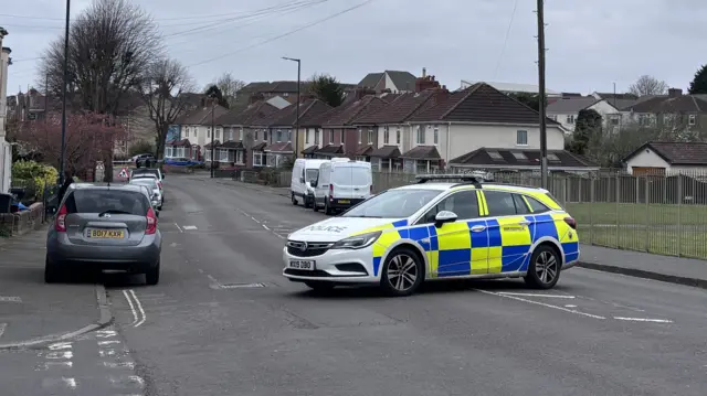 A police car blocks a road. A playing field is on the right side of the image, with rows of houses in the background. Parked cars line the street.