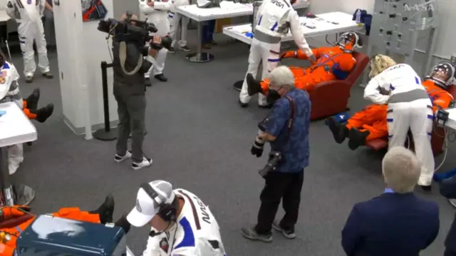 Two astronauts can be seen lying in a seat while Nasa workers adjust their suits