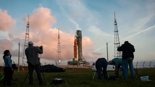 People with large cameras film the rocket, which is in the distance behind them