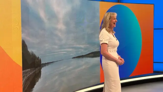 Carol pictured side on as she smiles standing in the Breakfast studio in front of a picture of a lake