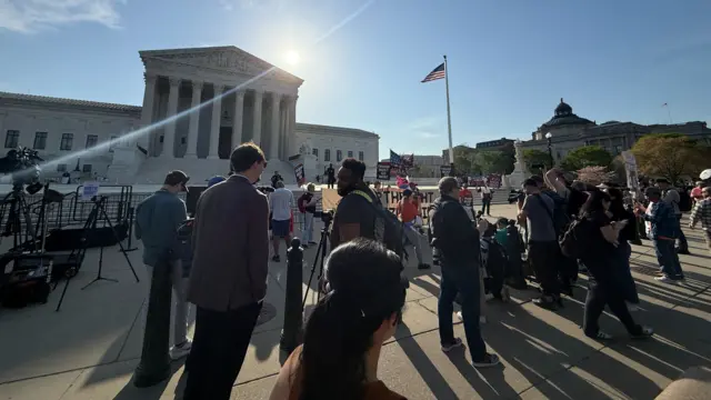 People gather outside the Supreme Court. Some hold signs, others have cameras set up. The building can be seen in the background