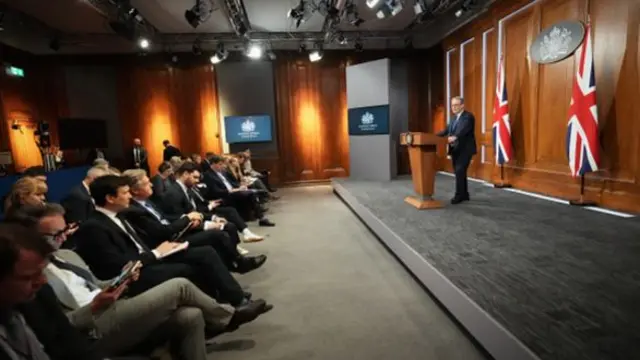A wide, side view of the press conference with Keir Starmer on the stage and the press sitting in seats.