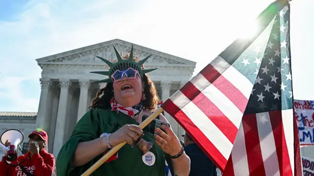 Протестующий в костюме Леди Свободы держит флаг США.