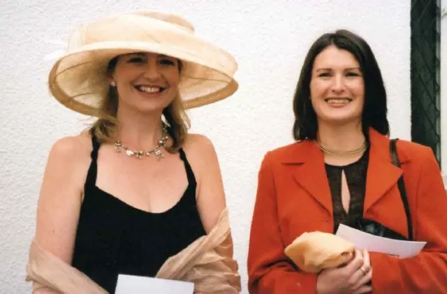 Carol and Helen smile at the camera standing next to one another. Each holds a bit of paper. They are dressed smartly and Carol wears a large hat