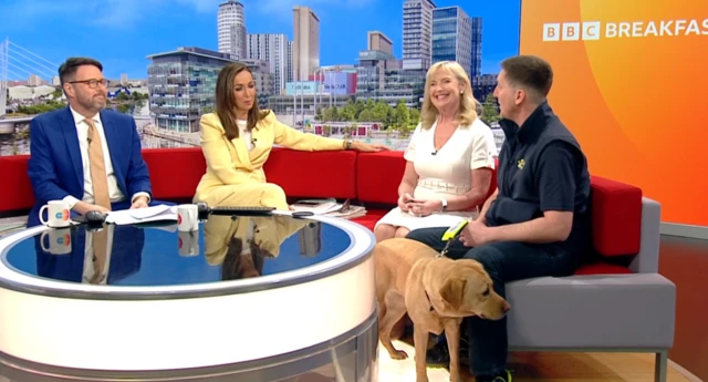 Carol sat next to a man on a sofa who holds a golden labrador on a lead while the presenters look on at them