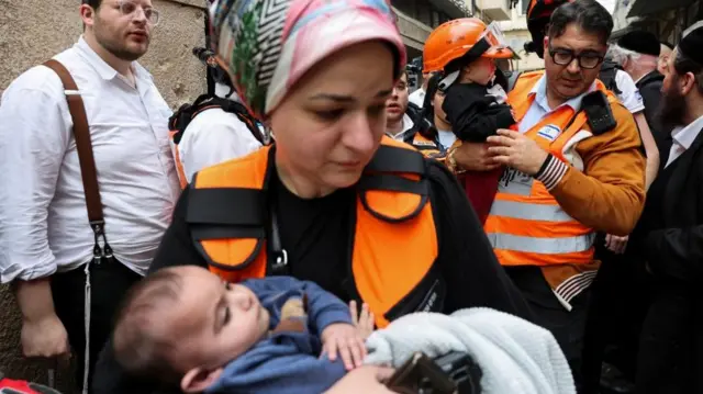 A female emergency worker wearing a black top, orange vest and coloured headscarf carries a baby wrapped in a blanket. Behind her, a male emergency worker also dressed in an orange vest carries another child - the child is wearing an orange safety helmet.