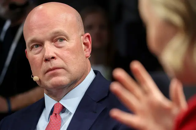Close up photo of Matthew Whitaker, US ambassador to NATO. He is wearing a suit ober a light blue shirt and a red tie. he is also wearing a brown microphone headset.