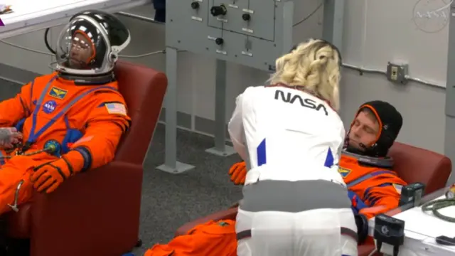 Two astronauts are seated - both wear orange suits and a woman in white is adjusting something on one of the astronaut's suits
