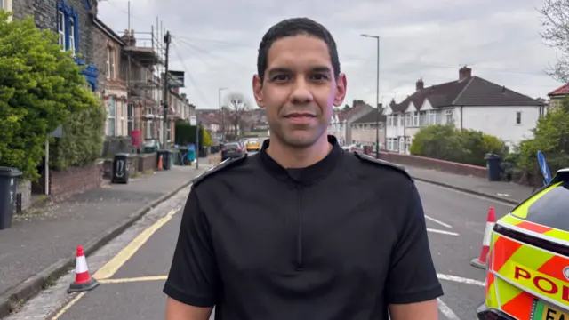 A police officer is seen from his shoulders up standing on a road.