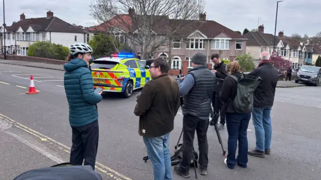 People gather around a parked police car on a residential road.