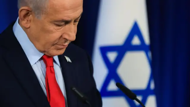 Israeli Prime Minister Benjamin Netanyahu looks down as he speaks at a lectern with the national flag in the background.
