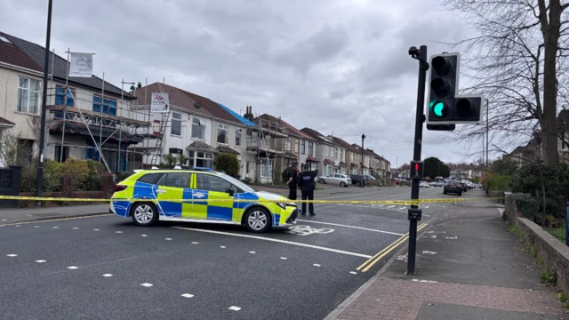A police car is parked in the middle of a road with a cordon around it and two officers on the road. There are residential houses either side.