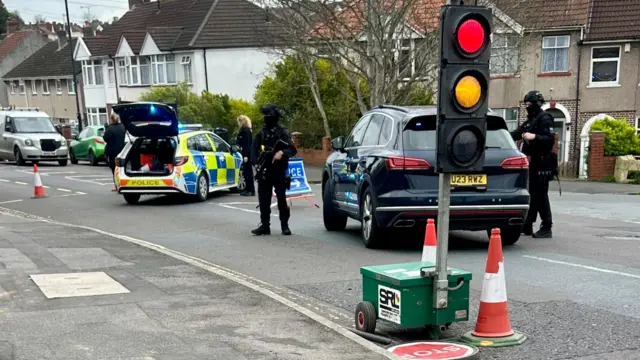 Armed police are standing next to two police cars by a temporary traffic light.