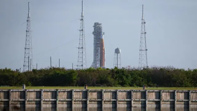 A far away view of the rocket which has multiple towers around it. There is a stretch of water, a field and tree cover in front of the rocket