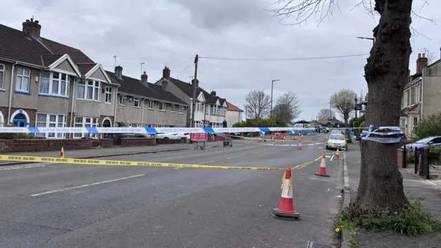 Two pieces of police tape are blocking off a section of a wide, residential street. There are cones lining the street.