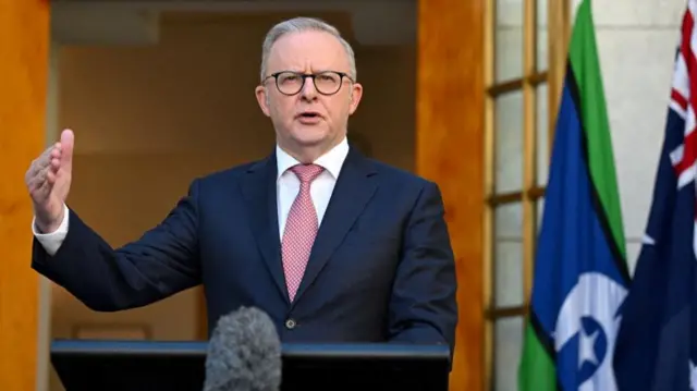 Anthony Albanese is shown speaking behind a lectern, with a microphone in the foreground. He is wearing a blue suit and white shirt with a pink tie.