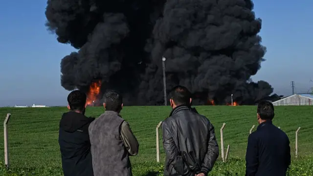 Four men with their backs to the camera, all dressed in dark clothing, stand watching a thick cloud of black smoke rising on the other side of a green field. Flames are visible at the base of the smoke.