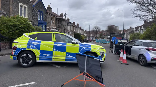 Police cars block road. One is blue and yellow and a second silver car to the right has its door open.