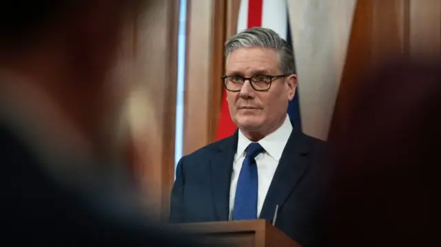 Keir Starmer mid-shot, picture taken from the back of the press room at No10 over the shoulders of members of the media. Starmer is standing at a podium in a dark suit, white shirt and blue tie