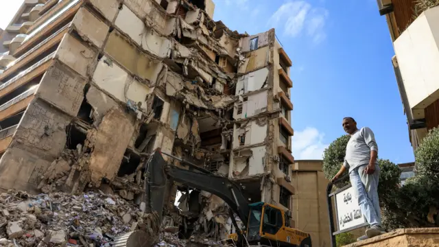 A man stands on rubble in Lebanon, with a digger behind him
