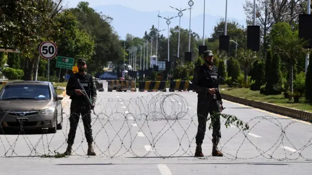 Two soldiers holding rifles stand in between rows of barbed wire in front of a blockade on an empty road