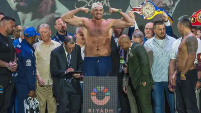 Tyson Fury holds up his arms as he weighs in with a crowd of people standing around him